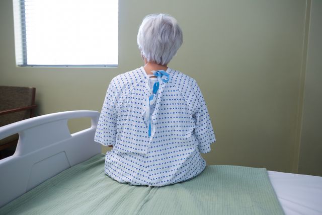 This image depicts a senior patient sitting on a hospital bed, facing away from the camera. The patient is wearing a hospital gown and appears to be in a pensive mood. The setting includes a window and a hospital bed, suggesting a clinical environment. This image can be used in healthcare-related content, articles about elderly care, medical treatment, patient experiences, and hospital settings.