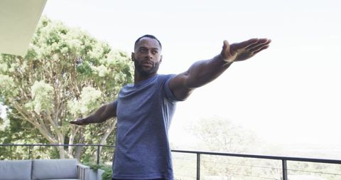 Man stretching arms on balcony wearing fitted gray t-shirt overlooking sunlit greenery
