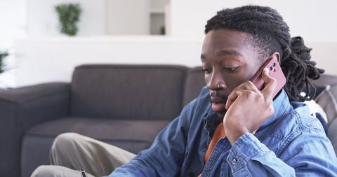 African american man working from home talking on phone and writing notes on sofa