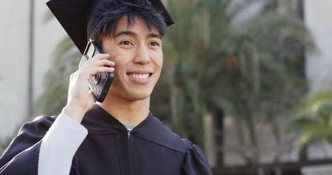 Smiling asian graduate talking on mobile phone in mortarboard and gown on campus