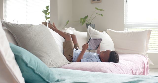 Young African American boy laying on a couch, engrossed in watching a tablet. The scene suggests a relaxed and comfortable home environment. The bright natural light and simple decor enhance the cozy atmosphere, portraying a blend of modern technology and home comfort. Ideal for use in materials related to technology for children, home life, comfortable living spaces, digital entertainment, and educational content for kids.