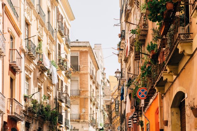 Narrow street bathed in sunlight features old buildings with balconies in a Mediterranean city. Clothes hanging on lines and plants on balconies add lively character to the urban scene. Ideal for marketing Europe travel destinations, urban architecture, cultural identity, and everyday street life.