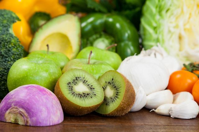 Close-up of a variety of fresh vegetables and fruits on a wooden table, including kiwi, green apples, garlic, avocado, broccoli, cabbage, turnip, and green pepper. Ideal for use in articles or advertisements about healthy eating, diet plans, organic produce, vegetarian and vegan lifestyles, and farm-to-table concepts.