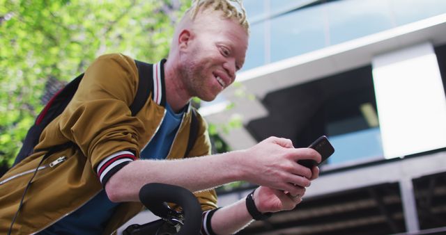 Young man in casual clothing smiling while using a smartphone near a bicycle. Urban environment suggesting an eco-friendly mode of transportation. Perfect for themes related to modern lifestyle, connectivity, transportation, and casual urban life.