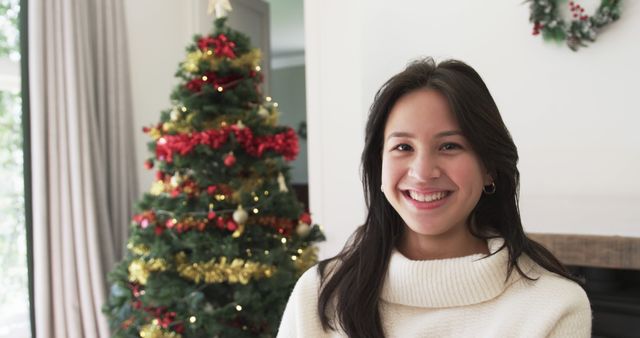 A young woman smiling while standing by a beautifully decorated Christmas tree indoors. The tree is adorned with red and gold decorations and twinkling lights, creating a festive atmosphere. Perfect for holiday-themed promotions, festive season marketing, Christmas greeting cards, and lifestyle blogs emphasizing holiday joy and indoor celebrations.
