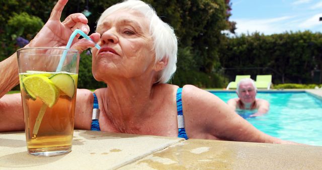 Senior woman enjoying a cold iced tea while lounging by a pool. An elderly man swimming in the background. Ideal for promoting senior lifestyles, summer leisure activities, retirement communities, and vacation advertisements.
