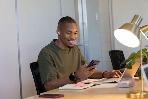 African american man smiling while working on smartphone and laptop at modern coworking
