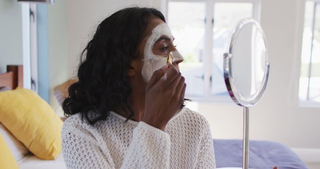 Woman applying facial mask in a well-lit room with windows. She is wearing casual clothing, indicating a relaxed and informal setting at home. This image can be used for promotional content related to skincare products, home self-care routines, health and wellness blogs, beauty tutorials, or lifestyle magazine features.
