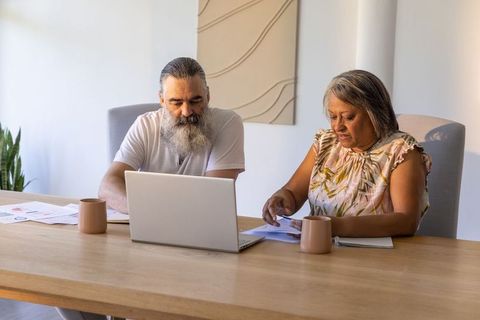 Man and senior woman reviewing documents on laptop at home dining table, planning finances