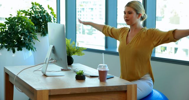 Woman balancing exercise and work while sitting on stability ball at desk. Demonstrates the importance of a healthy lifestyle and posture maintenance during work. Ideal for use in articles about workplace wellness, productivity, remote work setups, and ergonomic office solutions.