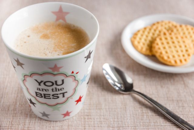 Coffee mug featuring motivational quote 'You are the Best' with light brown coffee, beside a plate with waffle biscuits and a spoon on a light-colored tablecloth. Ideal for promoting inspirational concepts, morning routines, beverages, breakfast snacks, positive thinking, and coffee culture.