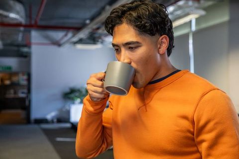 Young asian man sipping coffee while standing in modern industrial open office