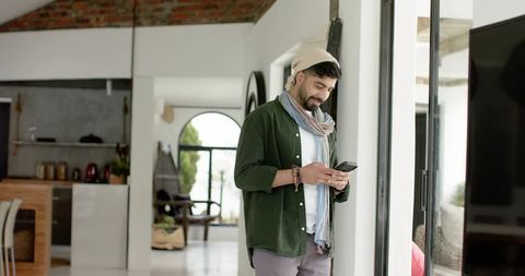 Indian man using smartphone by sliding glass door in modern loft interior, casual beanie and scarf