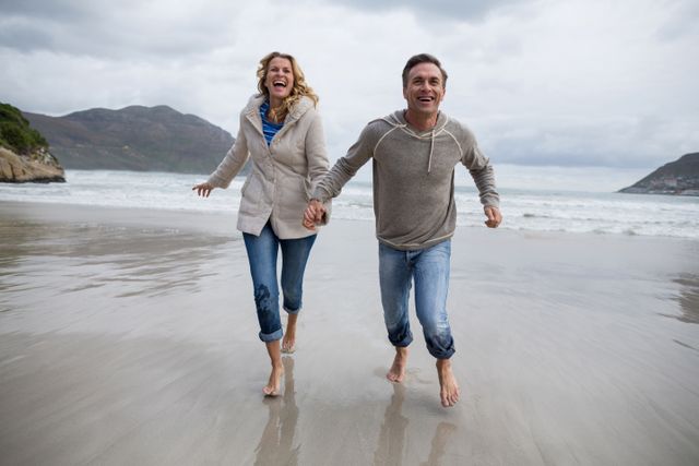 A joyful mature couple is running hand in hand along the beach, enjoying their time together. The ocean waves roll gently in the background with mountains and a cloudy sky completing the scenic view. Ideal for use in advertisements promoting outdoor activities, vacations, and romantic getaways. It is also perfect for lifestyle blogs, relationship articles, and health-focused content highlighting active lifestyles.
