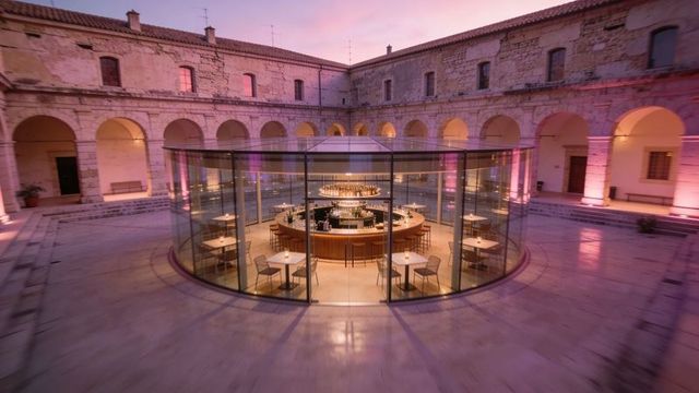 Glowing glass pavilion with circular wooden bar inside historic cloister at twilight