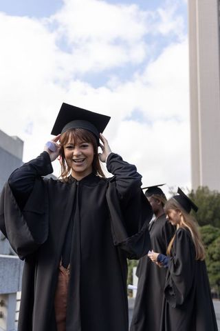 Young graduate smiling and adjusting mortarboard on campus terrace at commencement