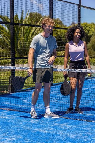 Mixed-race friends playing padel on vibrant blue court, holding black paddles, smiling outdoors