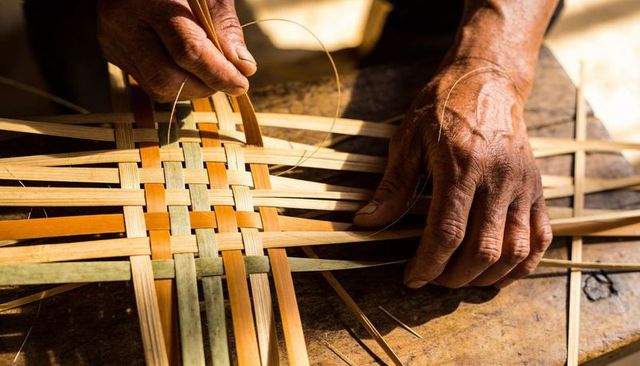 Elderly hands weaving multicolor bamboo reeds into woven basket lattice in sunlit workshop
