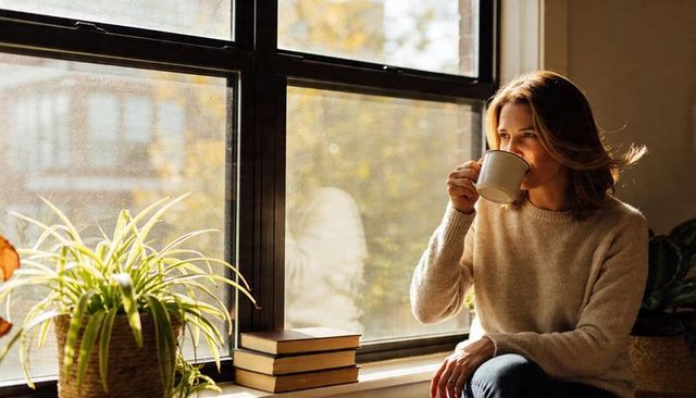 Woman sipping coffee on sunlit window ledge wearing cozy sweater and jeans, autumn morning