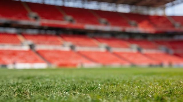 Dew-covered stadium turf gleaming in low-angle close-up with empty red stands and macro texture