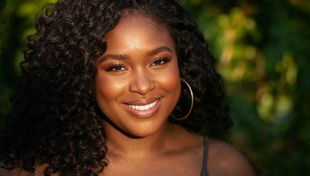 Smiling woman with curly hair wearing hoop earrings in sunlit garden headshot, golden hour