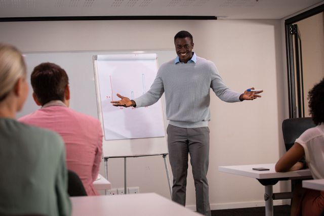 African American businessman smiling and gesturing while presenting to colleagues in a modern office. Ideal for use in business presentations, corporate training materials, teamwork and leadership articles, and office environment promotions.