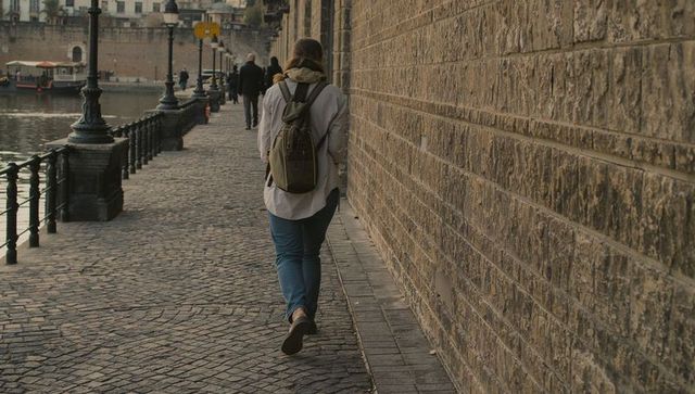 Walking woman on cobblestone riverside promenade wearing backpack and light coat