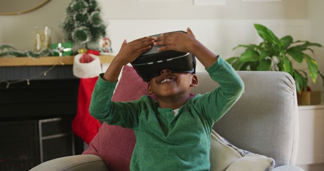 Boy wearing VR headset, smiling and looking excited, sitting on armchair in a cozy living room. Holiday decorations in background suggest festive atmosphere. Great for technology, family, holiday, and home-related themes, illustrating modern technology's impact on leisure activities.
