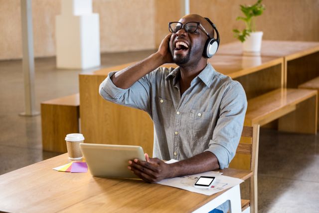 Man enjoying music while working on digital tablet in a modern office. Ideal for use in advertisements for technology products, office environments, or lifestyle blogs. Can be used to depict a relaxed and happy work atmosphere.