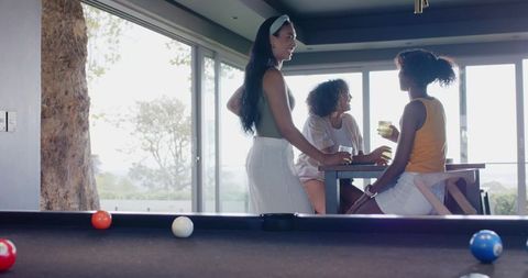 Three african american women enjoying drinks and chatting by pool table in modern lounge