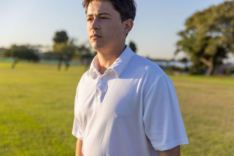 Young man standing and looking across sunlit park wearing white polo during golden hour