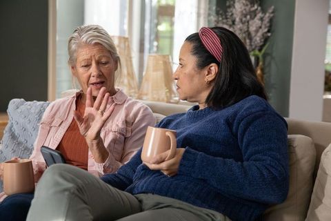 Two women chatting on cozy sofa sharing coffee and meaningful conversation in sunlit living room