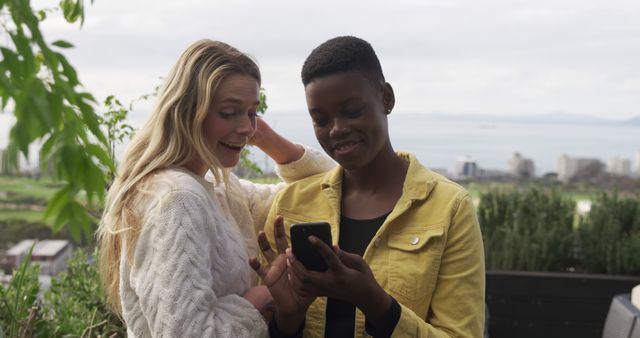 Two friends are outside, enjoying a pleasant day together while looking at a smartphone. They appear happy and engaged, standing against a backdrop of greenery and a scenic view of the city and water. Can be used for themes of friendship, technology, outdoor activities, leisure time, and diversity.