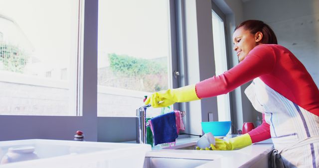 A young African American woman is cleaning a bathroom sink, wearing rubber gloves and an apron, with copy space. Her focused expression and the cleaning supplies suggest a thorough tidying up is underway.