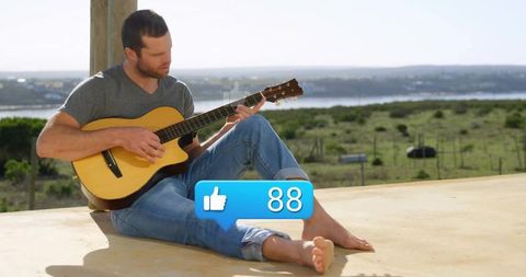 A man in a casual outfit relaxes on an outdoor veranda while playing an acoustic guitar. A blue thumbs-up icon with the number 88 hovers over him, representing social media engagement. The background reveals a picturesque and rustic setting with lush greenery, reflecting tranquility and the harmonious blend of modern and countryside life. This can be used to portray relaxation, bohemian lifestyle, or depict a social media theme intersecting with everyday leisure activities in serene environments.