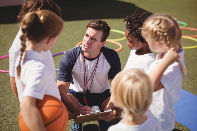 Coach is engaging with a group of children in a schoolyard, discussing possibly a sports strategy or game. The children are attentively listening, indicating a learning moment. This image can be used in educational materials, sports programs, team-building content, and advertisements for school activities or coaching services.