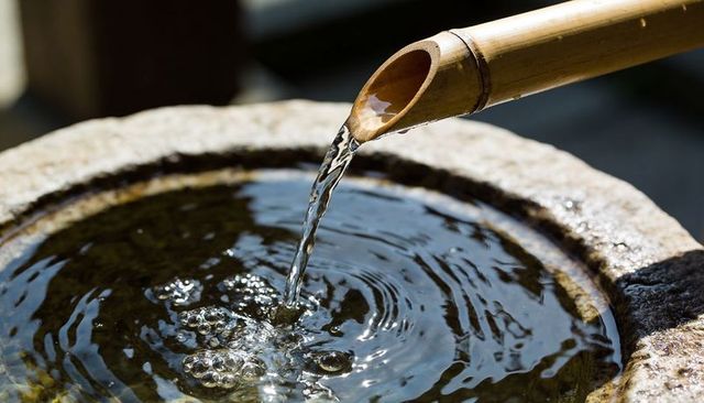 Sunlit bamboo spout pouring water into round stone basin creating ripples and bubbles