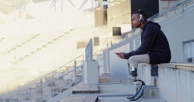 Man with prosthetic legs sitting on stadium steps listening to music. Ideal for use in sports, fitness, and motivation-related projects to promote inclusivity and determination.