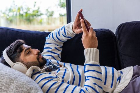 Young man reclining on sofa wearing headphones and striped sweater using smartphone