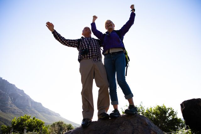 Low angle front view of a senior Caucasian couple enjoying time in nature together, hiking, taking a break, standing on a rock, holding hands up in a gesture of triumph