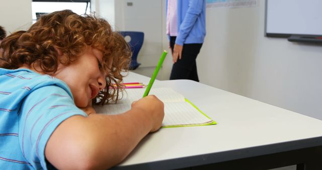 Young student falls asleep during class at desk, appearing tired while holding a pencil near an open notebook. Useful for illustrating academic stress, classroom fatigue, and the need for engagement in educational settings.