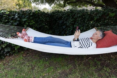 Mature woman relaxing on white hammock with smartphone and cozy socks in garden