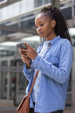 Young african woman standing outdoors checking smartphone carrying brown crossbody bag
