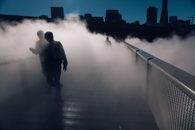 Silhouetted figures walking through dense fog on an urban bridge, with a city skyline in the background. This creates a mysterious and eerie atmosphere, perfect for use in projects related to urban themes, mystery genres, or atmospheric cityscapes.
