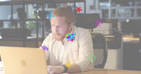 Focused red-haired man typing on laptop in creative office with colorful doodle overlays