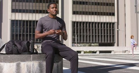 African American man sitting on concrete ledge checking phone with duffel bag in urban plaza