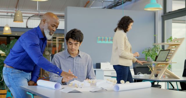 Architects collaborating in a modern office environment. One architect points to a building model while another discusses details. Ideal for business teamwork, corporate culture, architectural firms, and collaborative workspaces.