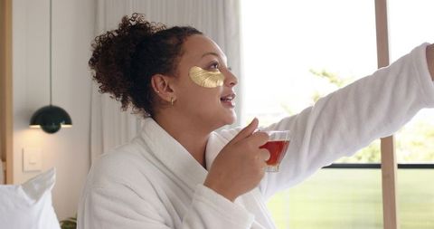 African american woman wearing gold eye patches holding tea in bathrobe morning routine