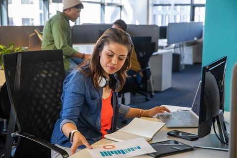 Young professional reviewing charts at open-plan desk with laptop and headphones