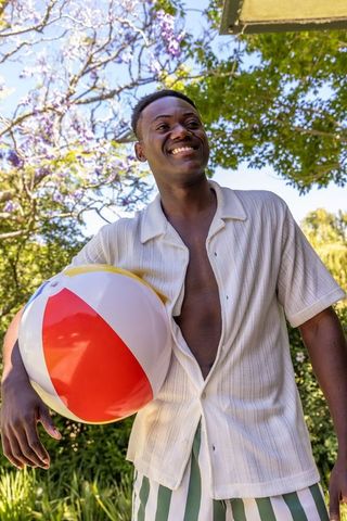 Smiling young man holding colorful beach ball in sunny backyard, wearing open shirt and swim trunks
